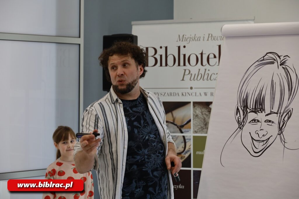 Na zdjęciu mężczyzna stojący przed flipchartem, trzymający flamaster, prawdopodobnie coś mówi. Nie patrzy na flipchart (na którym narysowana jest karykatura postaci), spogląda prawdopodobnie stronę słuchaczy (choć kadr ich nie obejmuje). Za mężczyzną stoi dziewczynka w wiekui  ok 7-9 lat.