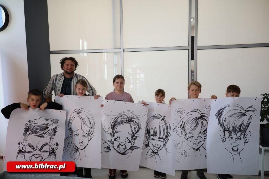 Na zdjęciu sześcioro dzieci trzymających duże (format ok A2/A1) rysunki - karykatury (rysowane czarnym na białym tle). Za dziećmi ustawiony mężczyzna (prawdopodobnie prowadzący zajęcia) - nieco dłuższe włosy, bródka, t-shirt i rozpięta koszula.