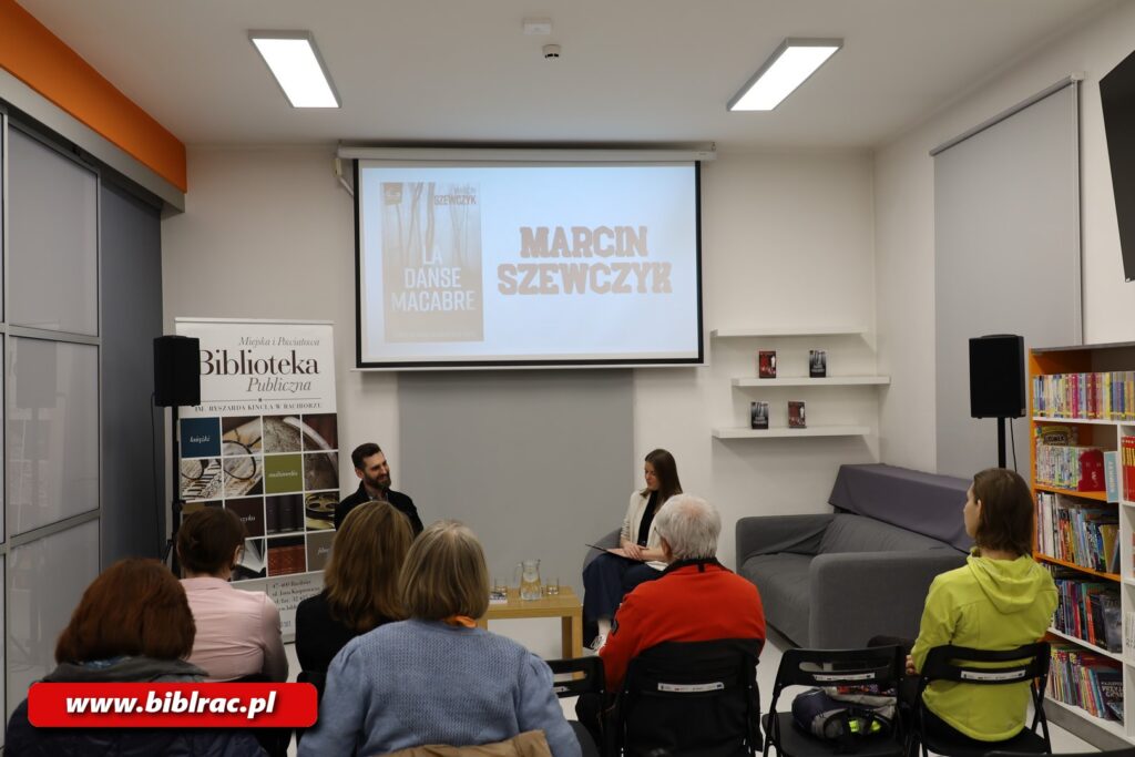 Na zdjęciu (kadr dosyć szeroki) siedzący w pomieszczeniu (prawdopodobnie bibliotecznym) mężczyzna śmiejący się podczas spotkania, obok niego siedząca kobieta pod ekranem (na który obraz rzuca projektor). Plecami do aparatu zwrócone są osoby siedzące na krzesłach (publiczność).