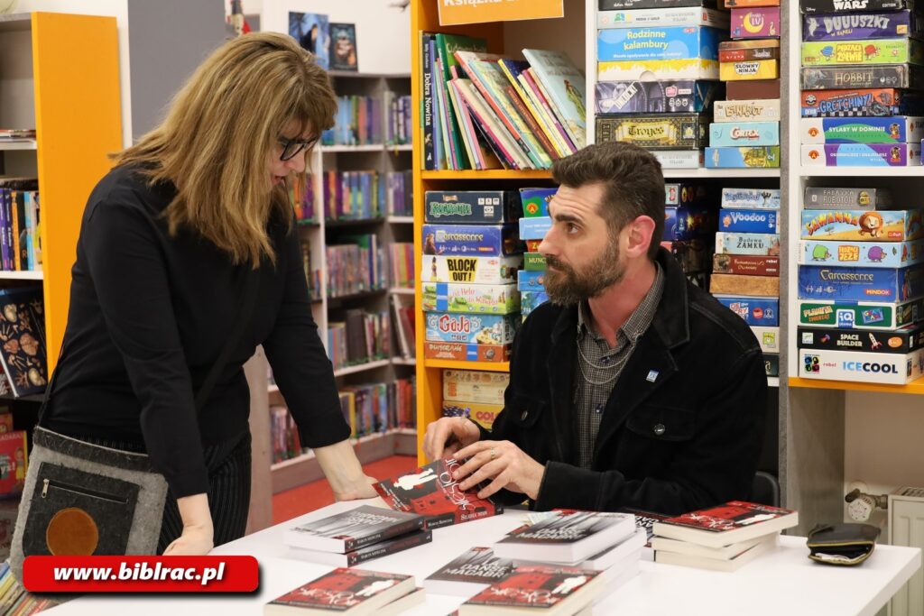 Na zdjęciu zbliżenie na kobietę (z lewej strony zdjęcia) stojącej i opierającej się rękami o stolik, przy którym siedzi mężczyzna z brodą. Mężczyzna spogląda na kobietę (ona na niego), otwiera lub zamyka leżącą na stoliku książkę (prawdopodobnie podpisywał ją lub będzie ją podpisywał).