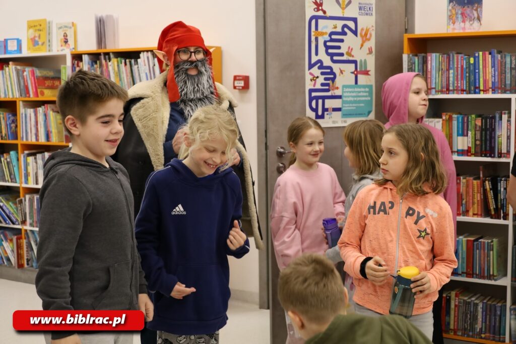 Na zdjęciu dzieci w pomieszczeniu z regałami i książkami, za nimi stoi kobieta przebrana za elfa (broda, sztuczne uszy, czapka. Dzieci są w wieku 6-11, niektóre się uśmiechają.