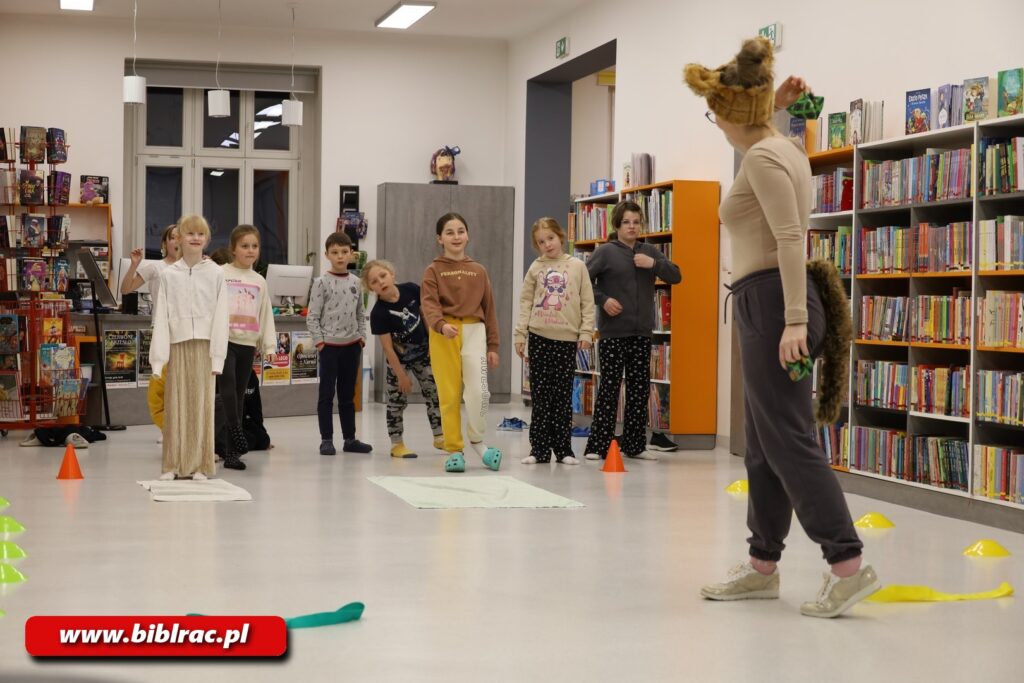 Na zdjęciu dzieci w wieku 6-10 lat stojące w rozproszeniu w pomieszczeniu z regałami i książkami. Dzieci są zwrócone przodem do aparatu, ale nie pozują, patrzą na dorosłą kobietę z dziwną futrzastą brązową opaską z uszami na głowie. Kobieta ta jest odwrócona prawie całkiem od aparatu, zwrócona jest natomiast w stronę dzieci, wygląda jakby dopiero zrobiła krok do przodu. Dzieci się uśmiechają, maja przed sobą na podłodze jakieś małe maty. Poza tym na podłodze w różnych miejscach rozłożone są małe pachołki i szarfy. Scenka rozgrywa się w pomieszczeniu, w tle widać regały z książkami.