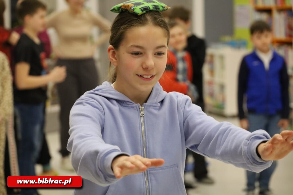 Na zdjęciu zbliżenie na dziewczynkę w wieku 9-12 lat, która ma wyciągnięte ręce do przodu, na głowie prawdopodobnie mały kwadratowy/prostokątny worek z wypełnieniem. W tle za dziewczynką sylwetki innych dzieci.