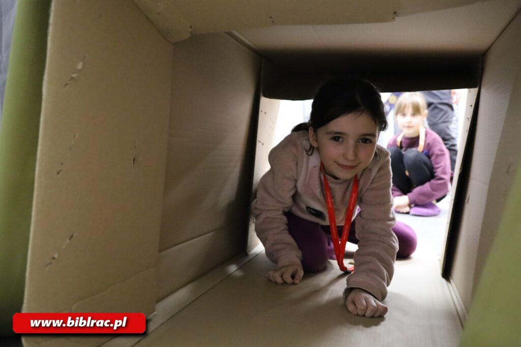 Na zdjęciu dziewczynka w wieku 7-10 lat (ciemne włosy, uśmiech, spogląda w stronę aparatu) idąca na czworakach przez tunek zrobiony prawdopodobnie na kartonów. Ścianki tego tunelu wypełniają dookoła kadr zdjęcia. W tle widać inną dziewczynkę, przed tunelem.