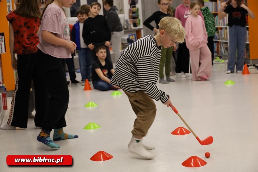 Na zdjęciu na pierwszym planie chłopak w wieku 8-12 lat sfotografowany od boku, pochylony, trzyma w rękach dziecięcy kij golfowy skierowany ku podłodze, na której widać małą pomarańczowa piłeczkę. Poza tym na podłodze porozstawiane są małe pachołki pomarańczowe i neonowo zielone. Chłopak ma blond włosy (z tylu podgolone, z przodu grzywka), za nim w kolejce stoją inne dzieci.
