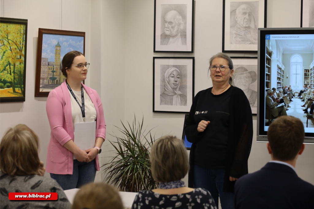 Na zdjęciu 2 kobiety stojące przed publiką w pomieszczeniu. Na ścianach obrazy i rysunki. Za wspomnianymi kobietami (jedna w szarym rozpinanym swetrze, druga ubrana na czarno) ekran wyświetlający jakiś slajd.
