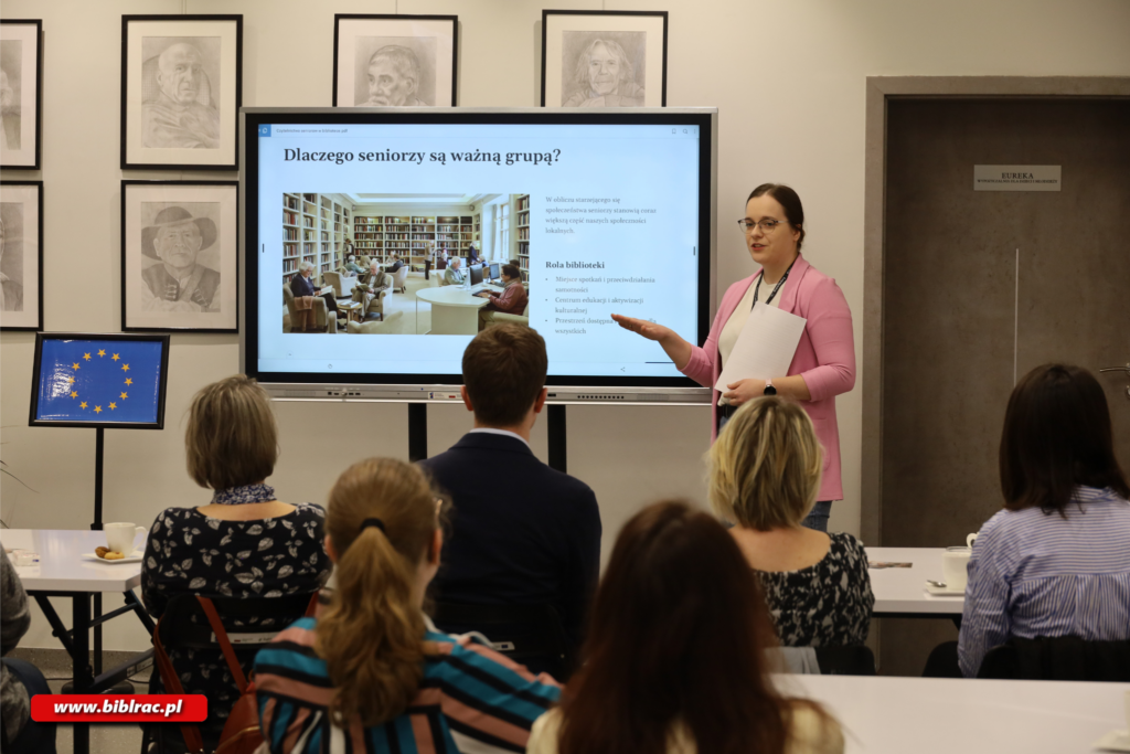Na zdjęciu kobieta w okularach w rozpiętym różowym swetrze przemawiająca do publiki, na ekranie za występująca kobietą wyświetlany jest slajd przedstawiający starsze osoby siedzące wygodnie w bibliotece. Kobiecie przysłuchują się osoby siedzące na krzesłach w pomieszczeniu, na którego ścianach wiszą obrazy i rysunki.