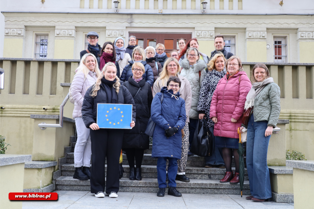 Na zdjęciu stojąca na schodach przed jasnożółtym zabytkowym budynkiem grupa kilkunastu osób, główne kobiet w różnym wieku oraz dwóch młodych mężczyzn. Ubrani w zimowe kurtki i płaszcze (śniegu brak). Jedna z kobiet, stojąca po lewej stronie u dołu, trzyma oprawiony w ramkę A3 wydruk flagi Unii Europejskiej. Grupa pozuje do zdjęcia, uśmiecha się.