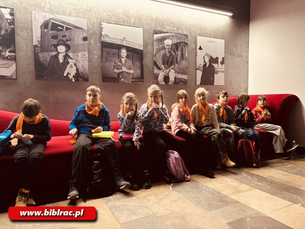 Na zdjęciu dzieci siedzące w holu na czerwonych siedziskach. Dzieci mają założone pomarańczowe husty na szyjach.