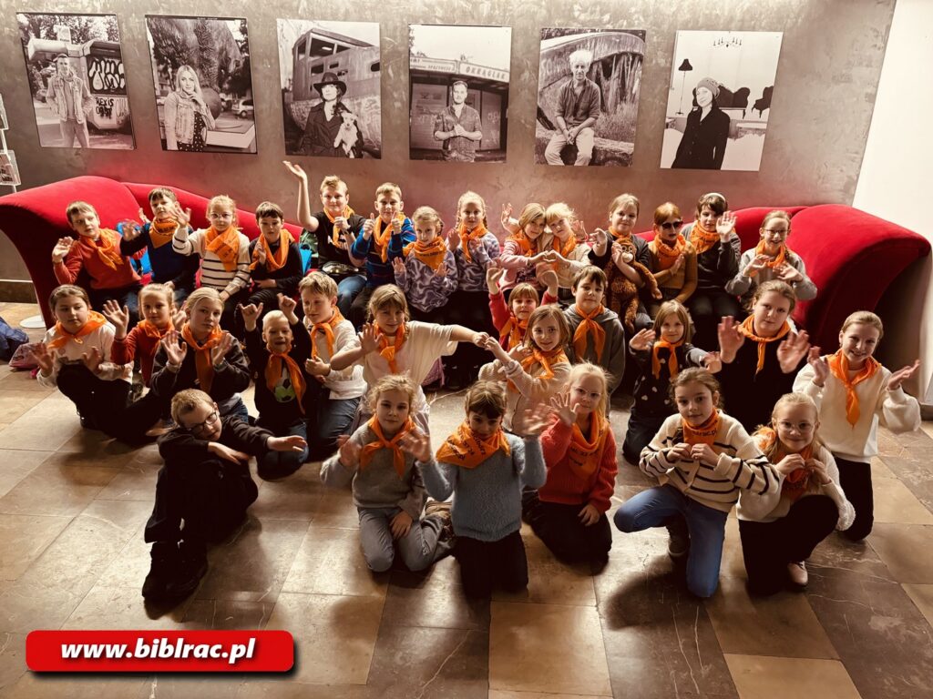 Na zdjeciu ok 20-30 dzieci siedzących na posadzce w holu. Na szyjach mają założone pomarańczowe chusty.