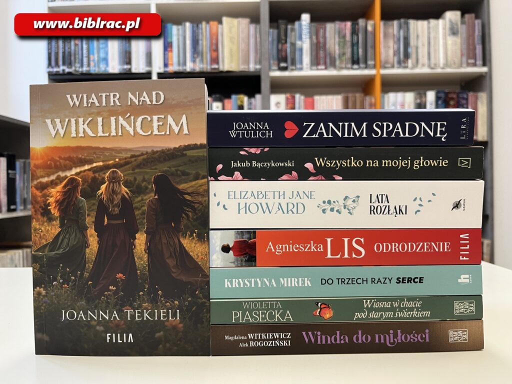 Na zdjęciu książki ułożone na stole - po prawej stronie stos książek zwróconych grzbietem do aparatu, po lewej stronie książka "Wiatr nad Wiklińcem" zwrócona okładką do aparatu. W tle regały z książkami.