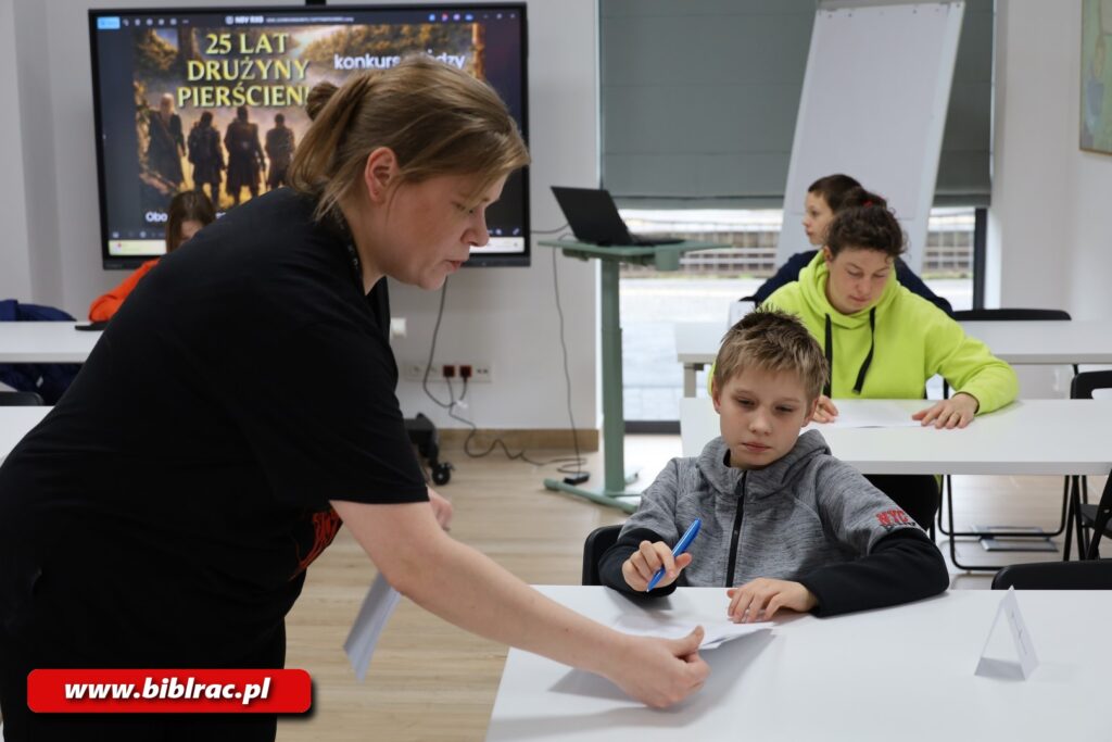 Na zdjęciu kobieta ustawiona bokiem do aparatu (niepozująca) kładąca na stole (przy którym siedzi chłopak w wieku 8-12 lat) kartki A4 (lub zabierająca je od chłopaka). Chłopak ma szaro-czarną bluzę (czarna jest dolna połowa, w tym rękawy od łokci do nadgarstków). Za chłopakiem są kolejne dwa stoliki i przy nich również siedzą dzieci. Z tyłu sali na ekranie wyświetlana jest grafika promująca konkurs wiedzy o Władcy Pierścieni.