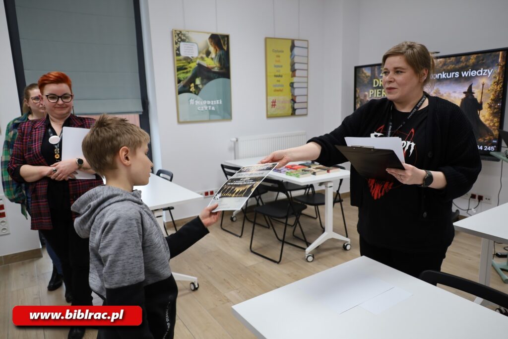 Na zdjęciu kobieta w czarnej bluzie i czarnych spodniach wręczająca dyplom chłopakowi w szaro-czarnej bluzie (chłopak w wieku 8-12 lat). Z lewej strony kadru inna kobieta obserwująca tę scenkę. W sali stoją białe stoliki, przy których nikt nie siedzi. Z tyłu sali na ekranie wyświetlana jest grafika promująca konkurs wiedzy o Władcy Pierścieni.
