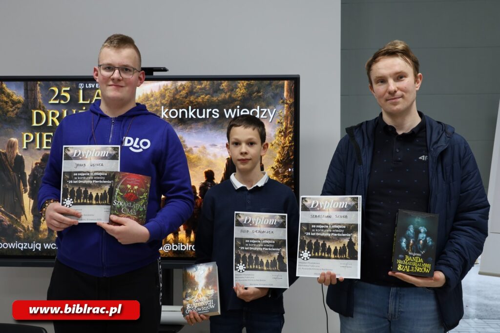 Na zdjęciu trzy osoby pozujące do zdjęcia, stojące w rzędzie - po lewej chłopak w wieku 15-19 lat (fioletowa bluza, okulary, jasne krótko ostrzyżone włosy trzyma w ręce dyplom oraz książkę, obok (w środku) sięgający mu do ramienia chłopczyk w wieku 9-12 lat, w sweterku granatowym z wystającym spod niego jasnym kołnierzykiem, czarne włosy, w rękach książka oraz dyplom, z prawej mężczyzna w wieku 25-32 lata, w rozpiętej wiosennej kurtce, włosy brąz. Wszyscy się uśmiechają, za nimi na ekranie wyświetla się grafika promująca konkurs wiedzy o Władcy Pierścieni.