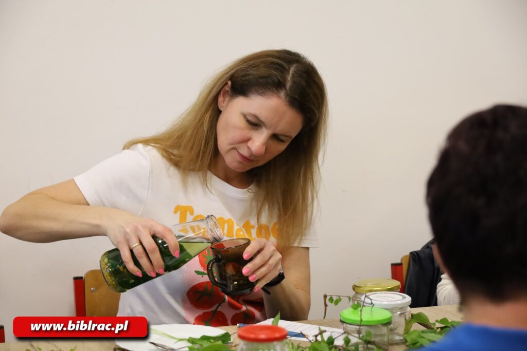 Zdjęcie przedstawia kobietę w długich blond włosach i białej bluzce z krótkim rękawem przelewającą jakiś płyn (w którym znajdują się zioła) z przezroczystej karafki / butelki do szklanki z ciemnego szkła. 