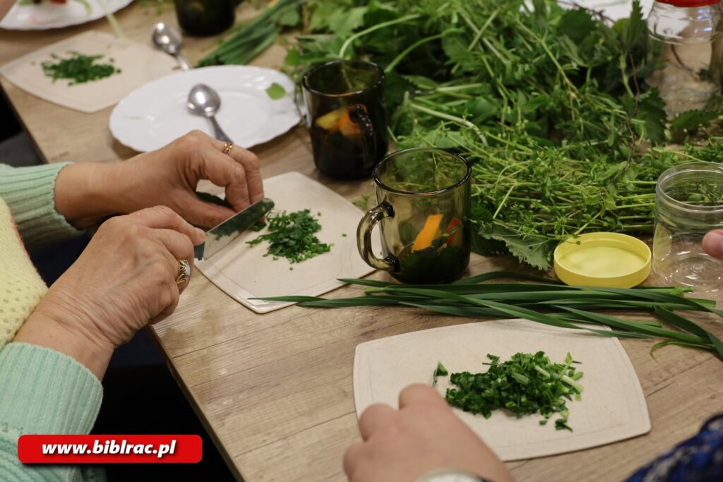 Na zdjęciu zbliżenie na stół, na którym znajdują się 2 podstawki z posiekanymi ziołami , a także szklanki z włożonymi kawałkami owoców, zioła leżące luźno ba stole, a także pusty talerzyk. Poza tym na widać też dłonie kobiet. Jedna z nich trzyma nóż, którym sieka zieleninę na tacce.