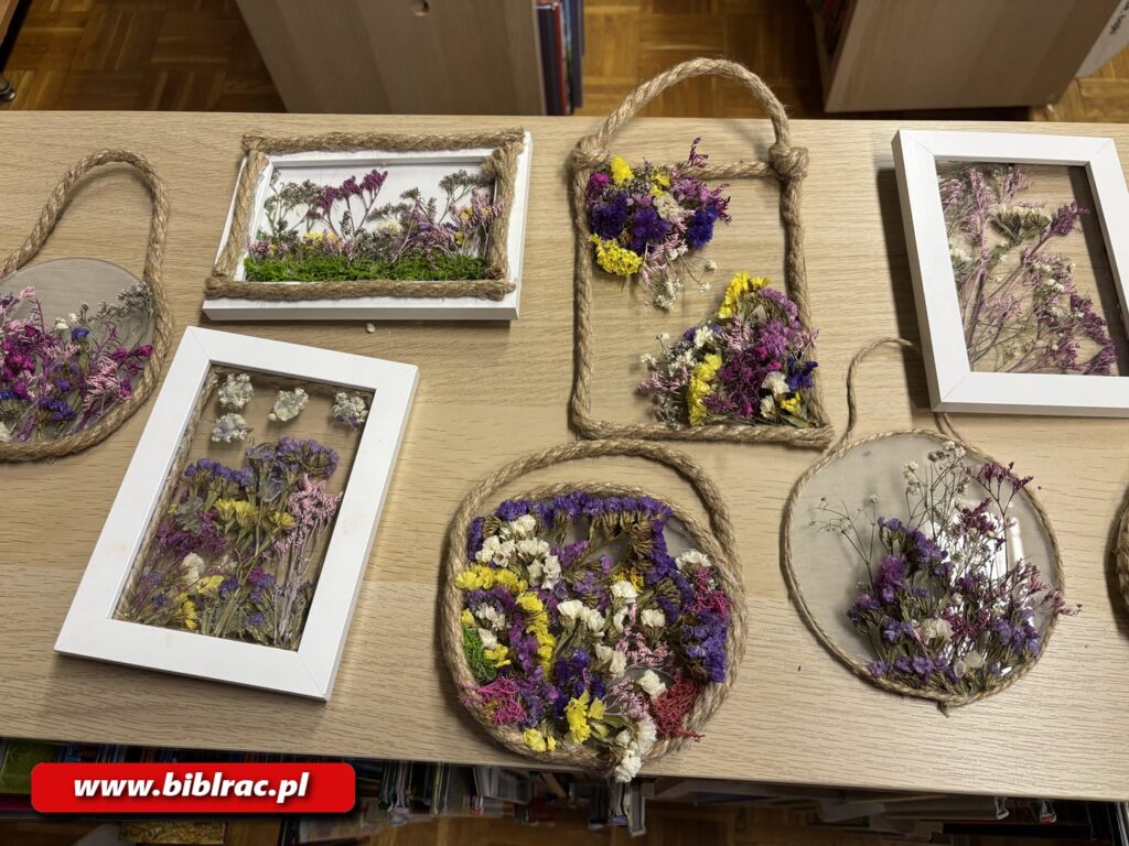 Na zdjęciu różne ozdoby z kwiatów ułożone na stole. Są to przede wszystkim kwiaty ułożone w tradycyjnych białych ramkach na zdjęcia (bez tła, bez "plecków"), oraz ułożone w okrągłych i prostokątnych ramkach wykonanych z grubszego sznurka (czasem z przezroczystym tłem z pleksi).