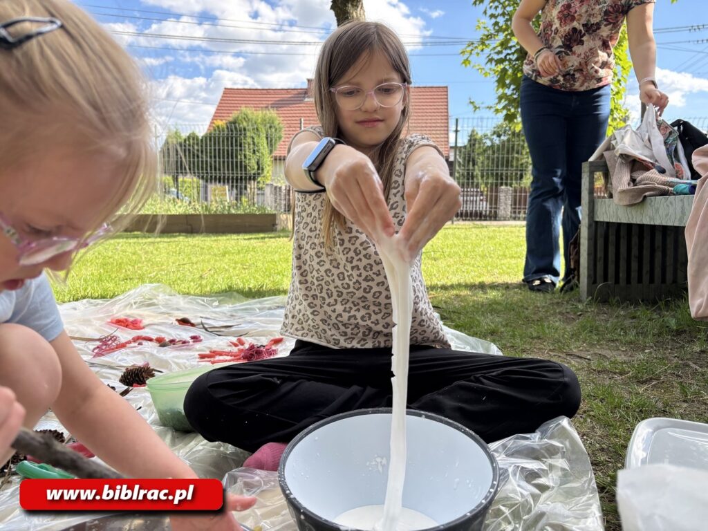 Na zdjęciu dziewczynka wlewająca jakąś białą płynną masę do miski. Dziwczynka siedzi na folii rozłożonej na trawniku. Widać też fragment twarzy innej dziewczynki siedzącej na pierwszym planie oraz fragment kobiety stojącej z tyłu poza folią. W tle płot, drzewa, dom.