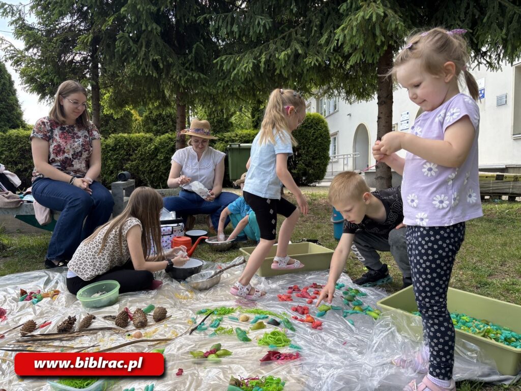 Na zdjęciu grupa dzieci w plenerze, układająca na rozłożonej folii swoje leśne znaleziska (szyszki itd.) oraz inne elementy (pomalowany makaron itd.), w tle dwie siedzące / kucające kobiety - jedna w okularach, druga w jasnym letnik kapeluszu. Na pierwsyzm planie dziewczynka w wieku 4-6 lat stojąca i oglądająca coś małego w dłoni