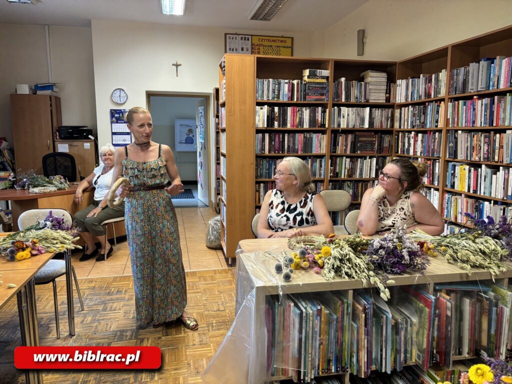 Na zdjęciu pomieszczenie biblioteczne, przy stole 2 kobiety tworzące wianki z kwiatów, obok stojąca kobieta w długiej sukni.