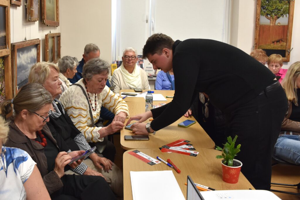 Na zdjęciu seniorzy siedzący przy stole. Poprzez stół nachyla się mężczyzna w czarnej koszuli i pokazujący coś na smartfonie jednej kobiecie siedzącej przy stole.