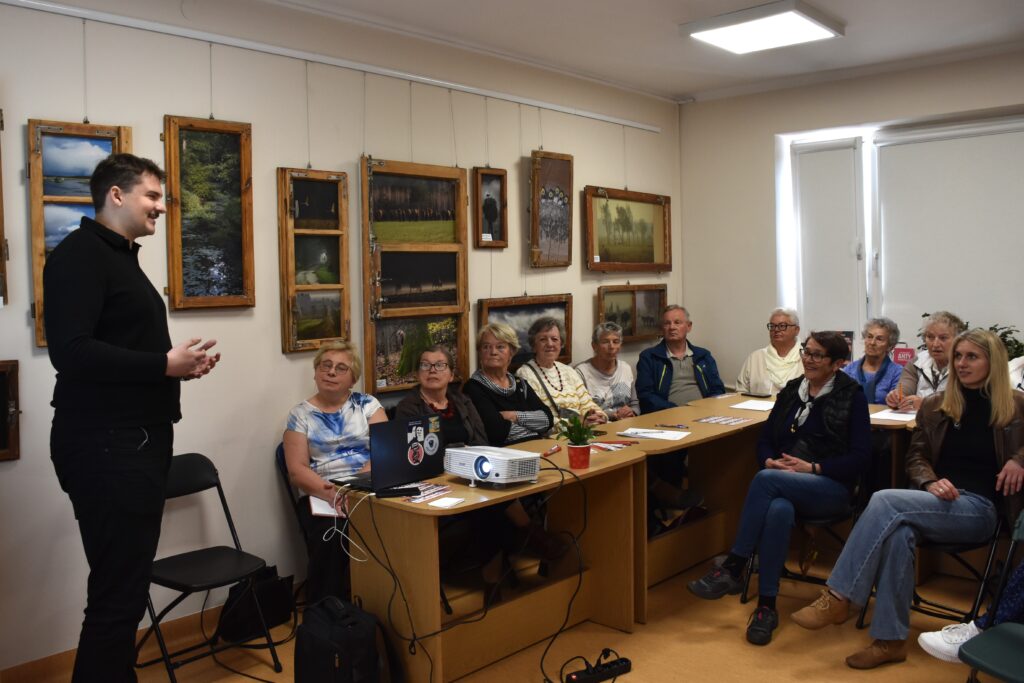 Na zdjęciu mężczyzna w czarnej koszuli stojący na środku sali przed seniorami (oraz młodszymi osobami) siedzącymi między innymi przy stołach.
