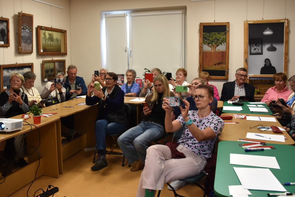 Na zdjęciu seniorzy oraz młodsze osoby siedzące przy stołach (oraz na krzesłach bez stołów) podnoszące do góry smartfony (jakby robiły zdjęcia aparatem wbudowanym w telefon).