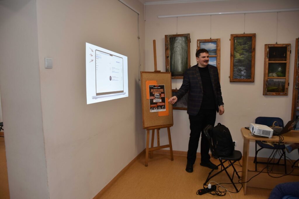 Na zdjęciu mężczyzna ubrany w czarną koszulkę, czarne spodnie i szarą marynarkę, z wąsem, uśmiechający się, pokazujący coś na obrazie wyświetlanym na ścianie przez rzutnik. Z anim ściana z obrazami.