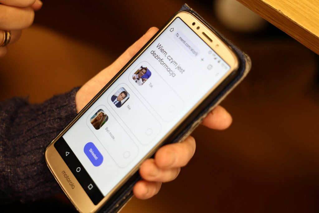 Na zdjęciu smartfon trzymany w dłoni, zdjecie zrobione od góry, na ekranie widać test z pytanie "Wiem czym jest dezinformacja" oraz odpowiedzi do wyboru: "tak", "nie", "być może". Obok odpowiedzi śmieszne obrazki z memów (zdjęcia z różnymi minami).