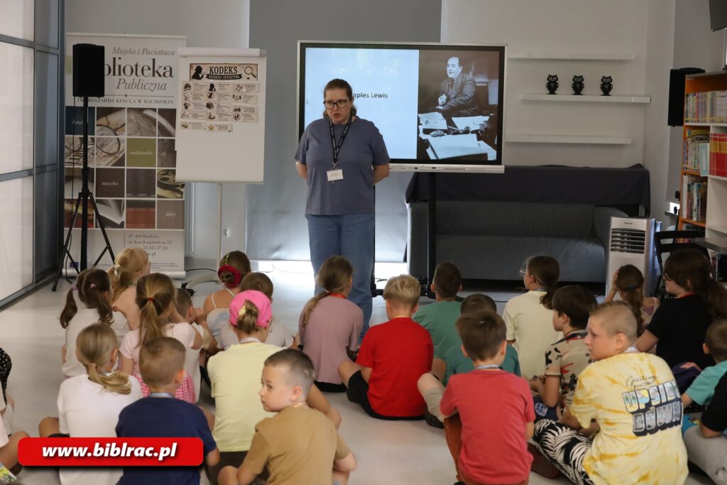 Na zdjęciu dziec w pomieszczeniu bibliotecznym słuchające stojącej przed nimi kobiety