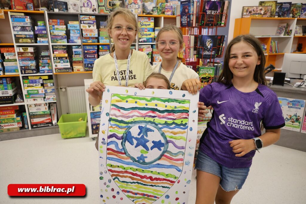 Na zdjęciu troje dzieci w wieku 7-12 lat w pomieszczeniu bibliotecznym trzymające wykonany ręcznie herb