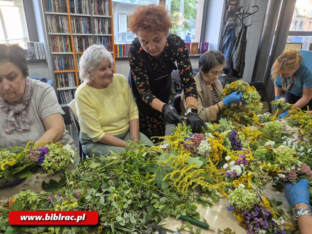 Na zdjęciu stół, na którym rozłożone są sterty roślin, przy stole siedzi 5 kobiet robiących wianki (jedna w zasadzie stoi). W tle regał z książkami.