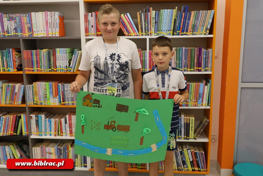 Na zdjęciu pozuje dwóch chłopaków, jeden w wieku 6-7 lat, drugi (wyższy, po lewej, uśmiechnięty) 9-12 lat. Obaj trzymają w pionie wykonaną papierową pracę 2D (duży arkusz zielonego brystolu a na nim przyklejony papierowy traktor, niebieska droga lub rzeka, drzewa, domy itd.).