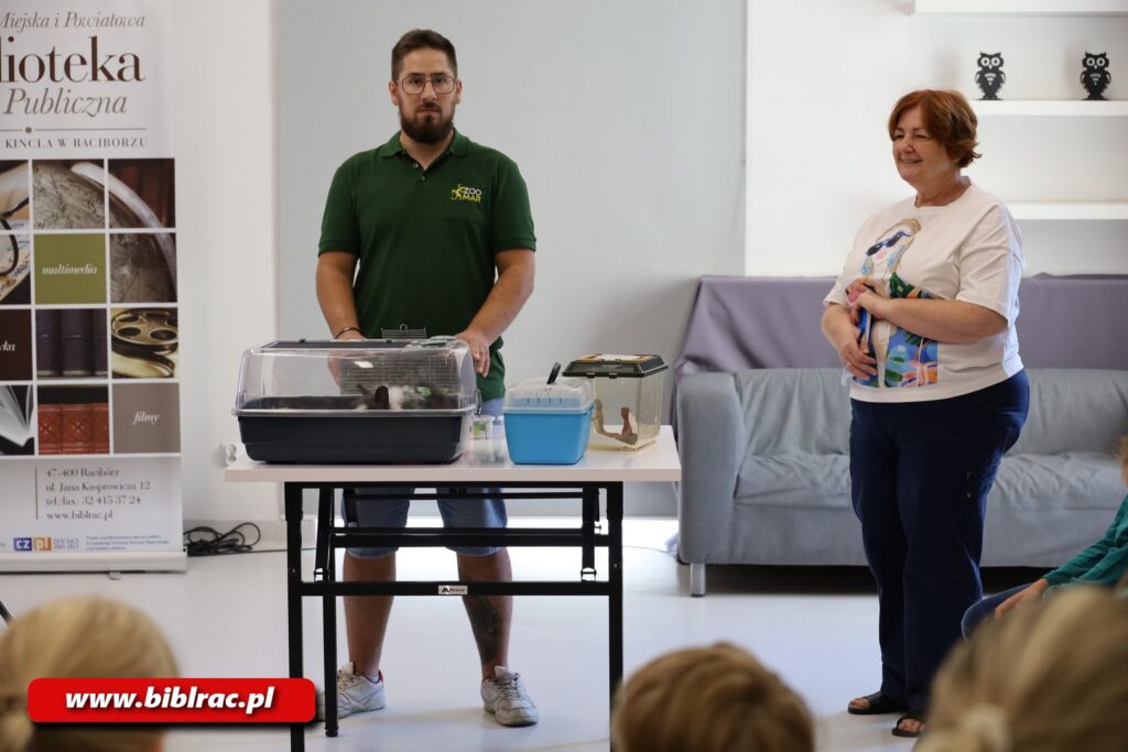 Na zdjęciu o szeroki kadrze męzczyzna w zielonej koszulce stojace za stołem z klatką dla zwierzątek (w środku królik) oraz przenośnym akwarium dla gadów (w środku gekon?), obok stojąca kobieta.