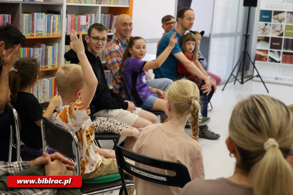 Na zdjęciu w pomieszczneiu bibliotecznym grupa dzieci siedzących na krzesłach (niektóre się zgłaszają). Kilkoro siedzi w towarzystwie mężczyzn (prawdopodobnie ojców).