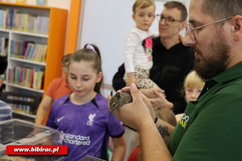 Na zdjęciu mężczyzna w zielonej koszulce trzymający na rękach koszatniczkę. Nieco dalej dziewczynka w fioletowej koszulce w wieku 10-12 lat. Jeszcze dalej mężczyzna w okularach i w czarnej koszulce trzymający na rękach chłopczyka w białej bluzie.