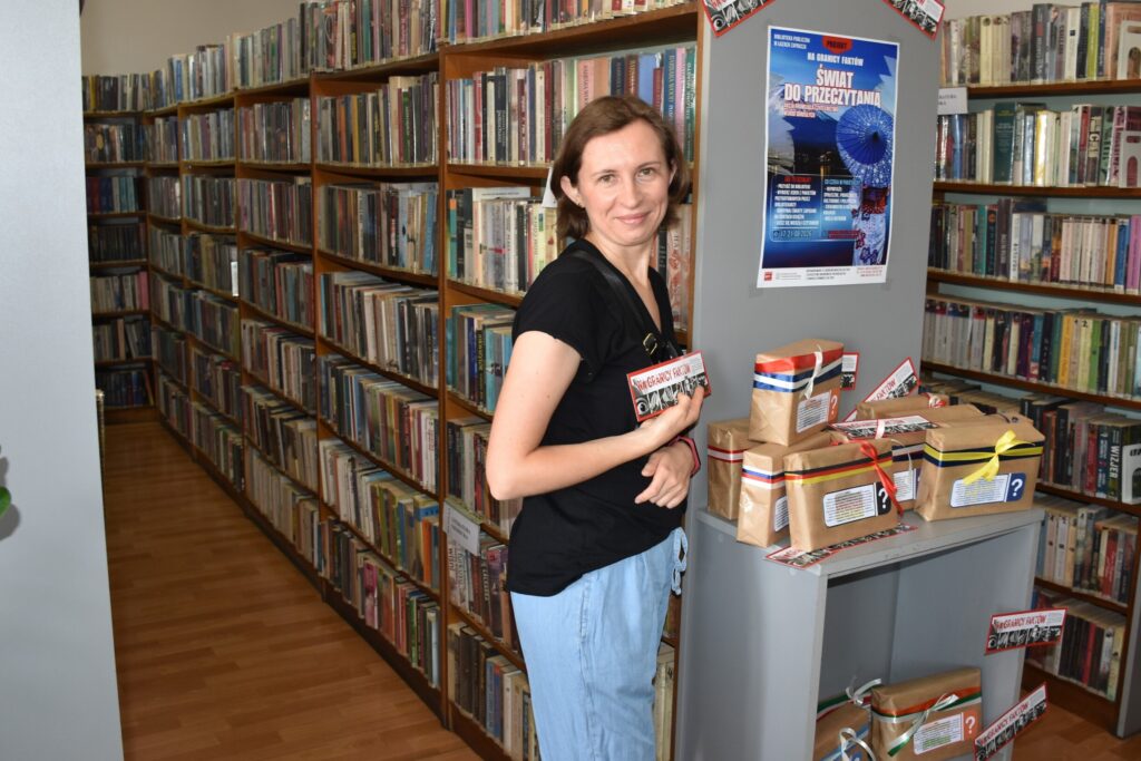 Na zdjęciu kobieta pozująca w bibliotece, stojąc przed szafką z pakunkami owiniętymi kolorowymi tasiemkami.