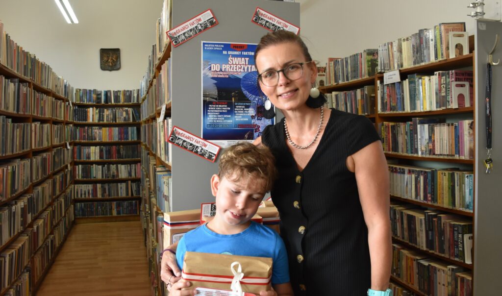 Na zdjęciu kobieta z dzieckiem (chłopcem kilkuletnim) pozująca w bibliotece, stojąc przed szafką z pakunkami owiniętymi kolorowymi tasiemkami.