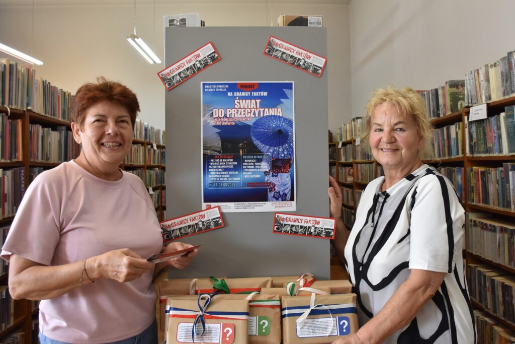 Na zdjęciu 2 kobiety pozujące w bibliotece, stojąc przed szafką z pakunkami owiniętymi kolorowymi tasiemkami.