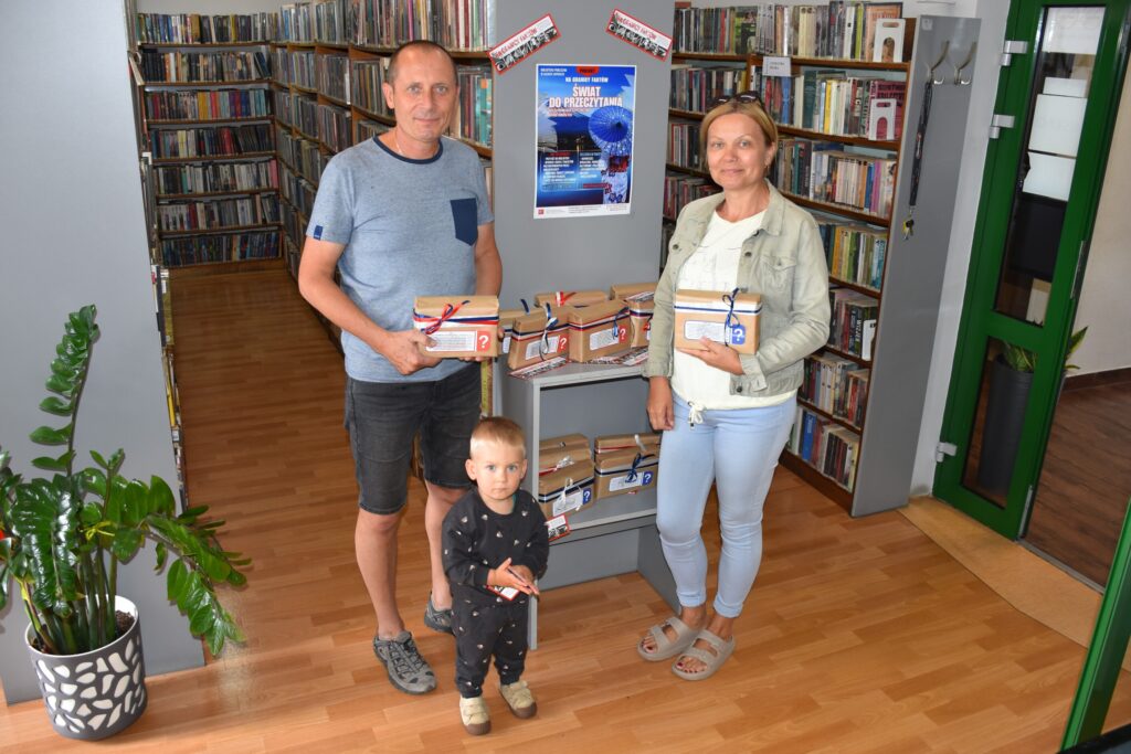 Na zdjęciu mężczyzna i kobieta z 1-2 letnim dzieckiem, pozujący w bibliotece, stojąc przed szafką z pakunkami owiniętymi kolorowymi tasiemkami.