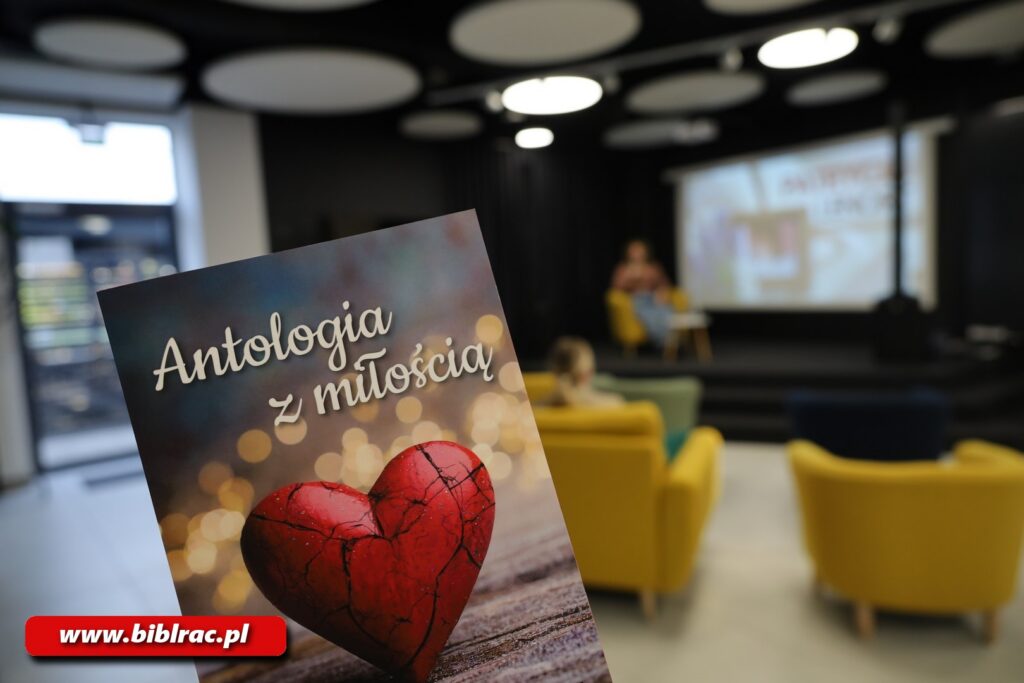 Na zdjęciu zbliżenie na okładkę książki Antologia z miłością (na okładce serce), w tle widać pomieszczenie ze sceną i żółtymi fotelami.